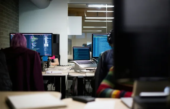 Two developers at adjacent desks working on monitors filled with lines of code in a dim office